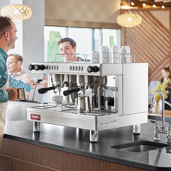 A man standing next to an Estella Caffe automatic espresso machine on a counter holding a cup of coffee.