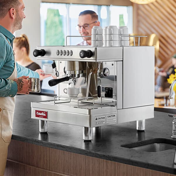 A man standing next to an Estella Caffe espresso machine on a counter.